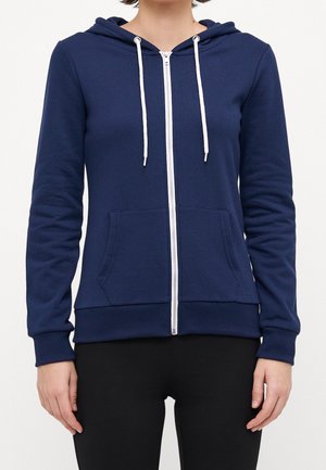 Vrouw draagt een marineblauwe hoodie met ritssluiting, witte trekkoorden en zakken aan de voorkant, gecombineerd met zwarte broek tegen een effen achtergrond.