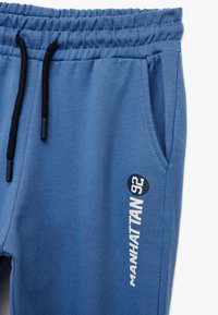 Blå sweatpants med elastisk talje, snor, sidelommer og "MANHATTAN 92" trykt i hvidt på venstre ben. Blødt stof.