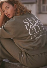 Tamsiai žaliai megzta maikutė su balta užrašu "SELF COUNTRY CLUB EST 2021" ir atitinkamos kelnės su mažu logotipu ant kojos.