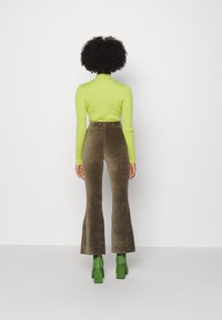 Haut vert à col roulé côtelé associé à un pantalon évasé en velours côtelé marron et des bottines vertes à talons blocs, vu de dos sur un fond uni.