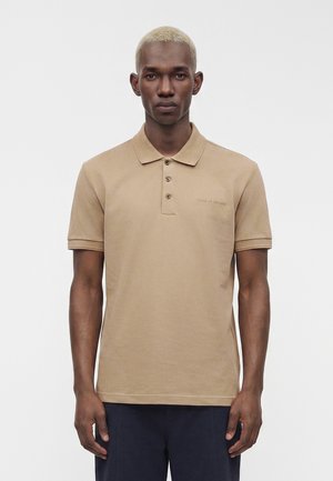Mann med kort blondt hår iført en beige kortermet poloshirt og mørke bukser, stående foran en ensfarget hvit bakgrunn.