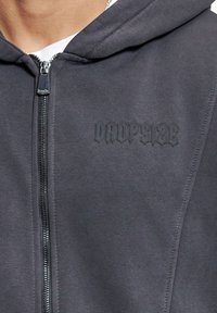 Sweat à capuche gris zippé en tissu doux, arborant un logo noir en relief "DROPSIDE" sur la poitrine et une fermeture éclair métallique.