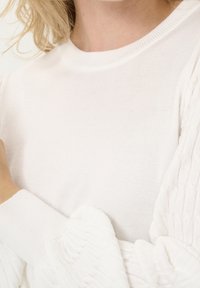 Vit tröja med rund halsringning och texturerade, volangärmar. Tyget verkar mjukt och ribbat med en slät kropp.