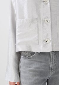 Silberne Satin-Cropped-Jacke mit strukturierter Oberfläche, ausgestattet mit vier Knöpfen und zwei Fronttaschen, kombiniert mit ausgebleichten grauen Jeans.