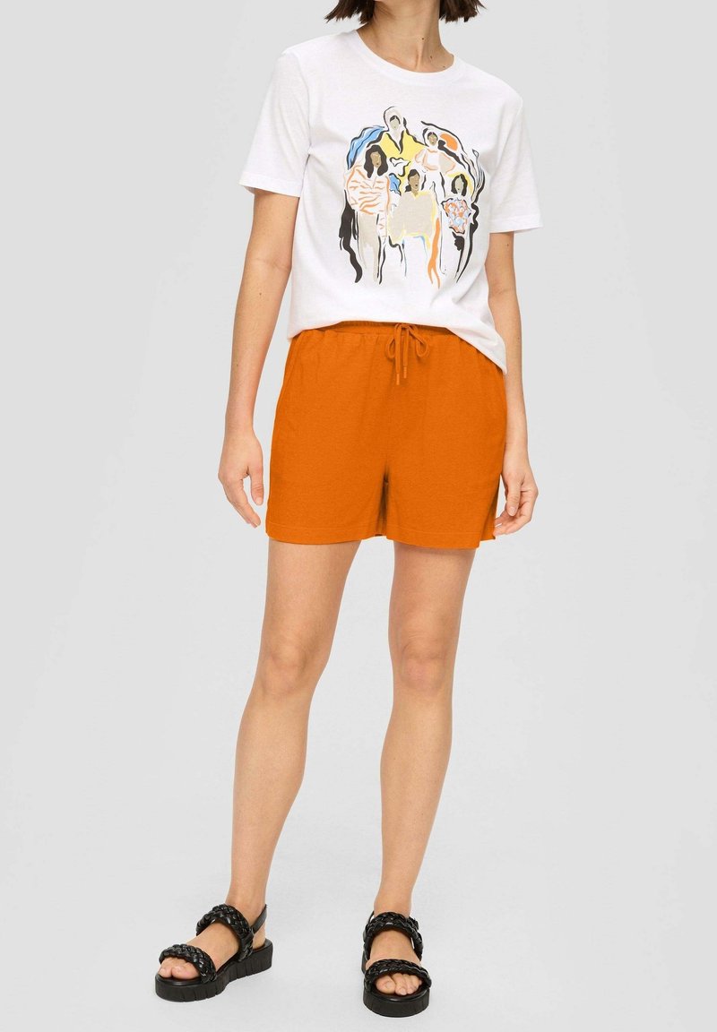 Femme portant un T-shirt blanc avec un imprimé abstrait multicolore de personnes, un short orange à cordon et des sandales noires tressées, debout devant un fond uni.
