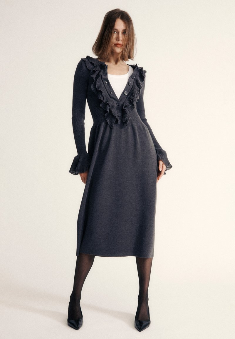 Abito in maglia grigio scuro con scollatura a volant, dettagli con bottoni, maniche lunghe, polsini a volant e texture liscia, abbinato a tacchi neri.