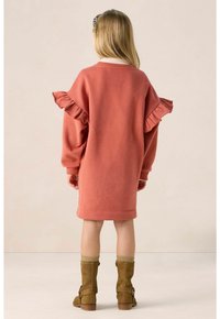 Rosa Sweatshirt-Kleid mit Rüschensäumen, Rundhalsausschnitt und geradem Saum. Getragen mit tanbraunen kniehohen Stiefeln und grünen gestrickten Socken.
