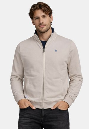 Mann mit kurzem braunem Haar, der eine hellbeige Reißverschlussjacke über einem dunkelblauen Hemd trägt, die Hände in den Taschen, vor einem schlichten Hintergrund stehend.