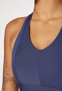 Mornarsko modra športna modrčka z rebrasto teksturo, širokimi naramnicami in V-vratnim dizajnom. Vključuje čiste šive in prilegajočo silhueto.