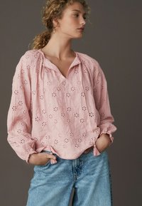 Blusa rosa ricamata con scollo a V, maniche lunghe, polsini arricciati e dettagli con frange, caratterizzata da motivi floreali e una vestibilità morbida.