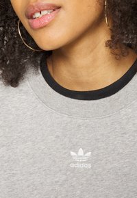 Felpa grigia chiara con colletto a coste nero. Presenta il logo Adidas ricamato in bianco sulla parte anteriore. Tessuto morbido e strutturato.