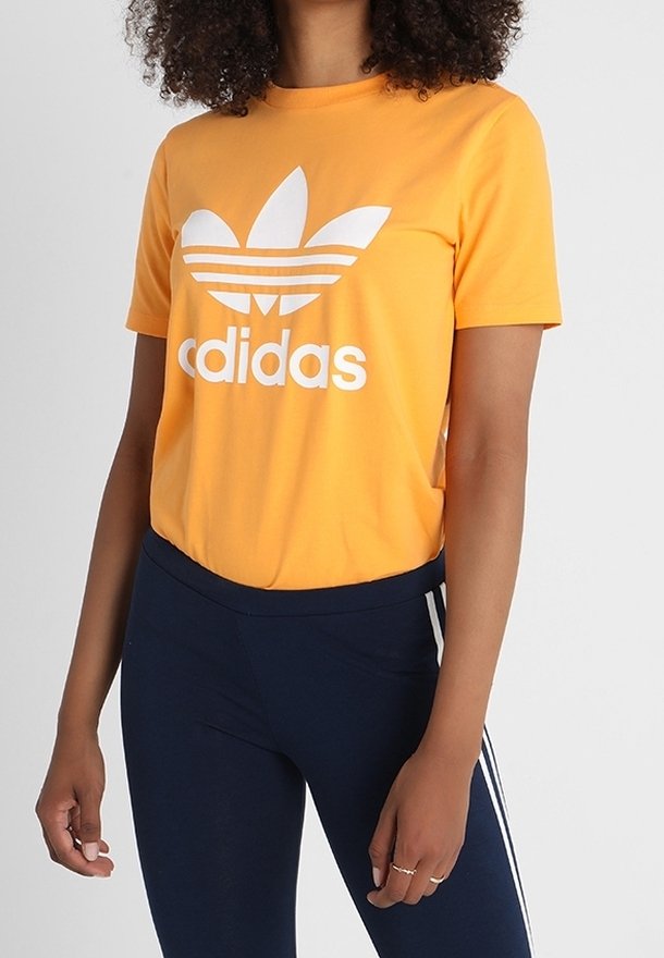 T-shirt en coton orange avec un logo Adidas blanc et trois rayures horizontales sur les manches. Associé à des leggings navy avec des rayures blanches sur les côtés.