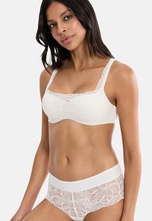 Kobieta w białym koronkowym biustonoszu z kwiatowym wzorem i pasujących koronkowych majtkach na jednolitym tle.