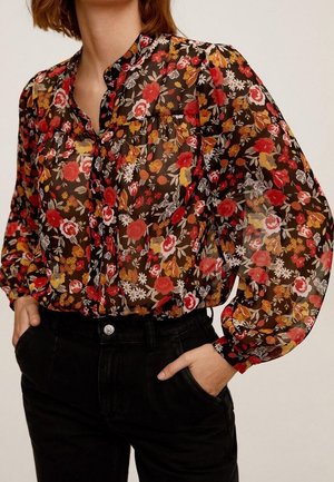 Žena v průsvitné černé blůze s červeným, oranžovým a bílým květinovým vzorem, v černých kalhotách, s rukama v kapsách, stojící před jednobarevným pozadím.