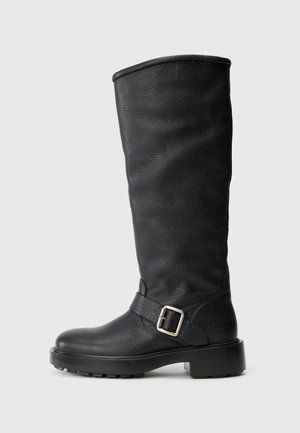 Bota preta de couro até ao joelho com acabamento texturizado, cano largo e detalhe de cinta com fivela. Apresenta uma sola grossa para maior aderência.
