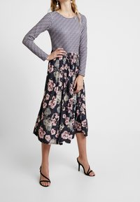 Vrouw in een midikleding met een geometrisch patroon aan de bovenkant en een bloemenprint rok, gecombineerd met zwarte hoge hakken.