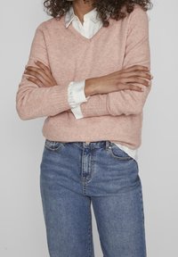 Une personne aux cheveux bouclés porte un pull-over V en rose clair sur une chemise à col blanc et un jean bleu, les bras croisés contre un fond gris.