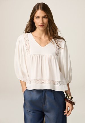 Witte blouse met een V-hals en kantdetails op de schouders en zoom. Zachte stof en ballonmouwen. Gecombineerd met denim broek.