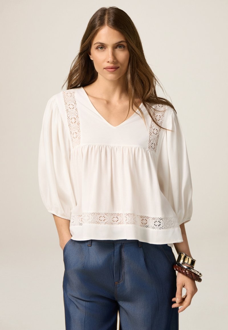 Weiße Bluse mit V-Ausschnitt und Spitzendetails an den Schultern und am Saum. Weicher Stoff und Ballonärmel. Kombiniert mit Jeans.