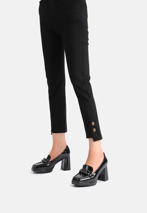 Sorte slim-fit ankelbukser med guldknapper nær manchetterne, båret med sorte laklæder loafers med blokhæle og kædedetalje.