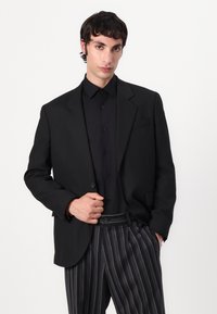 Black tailored blazer with notch lapels, single button closure, and two front pockets, paired with a black shirt and striped trousers.
