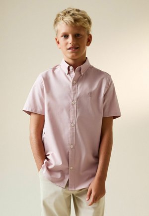 Băiat blond stând cu mâinile în buzunare, purtând o cămașă roz deschis cu mâneci scurte și nasturi și pantaloni deschiși la culoare, pe un fundal bej.