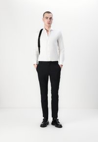 Weißes Hemd mit langen Ärmeln, kombiniert mit schwarzen Slim-Fit-Hosen und schwarzen Schuhen, akzentuiert durch einen schwarzen Gurt über der Schulter.