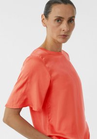Koraalkleurige blouse met korte mouwen van gladde stof, met een ronde halslijn en een losse pasvorm, benadrukt een effen kleur zonder patronen.