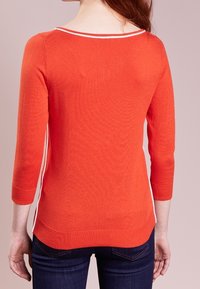 Pull en maille orange à manches trois-quarts, bordure blanche au niveau de l'encolure et des côtés, présentant une coupe ajustée et une texture lisse.