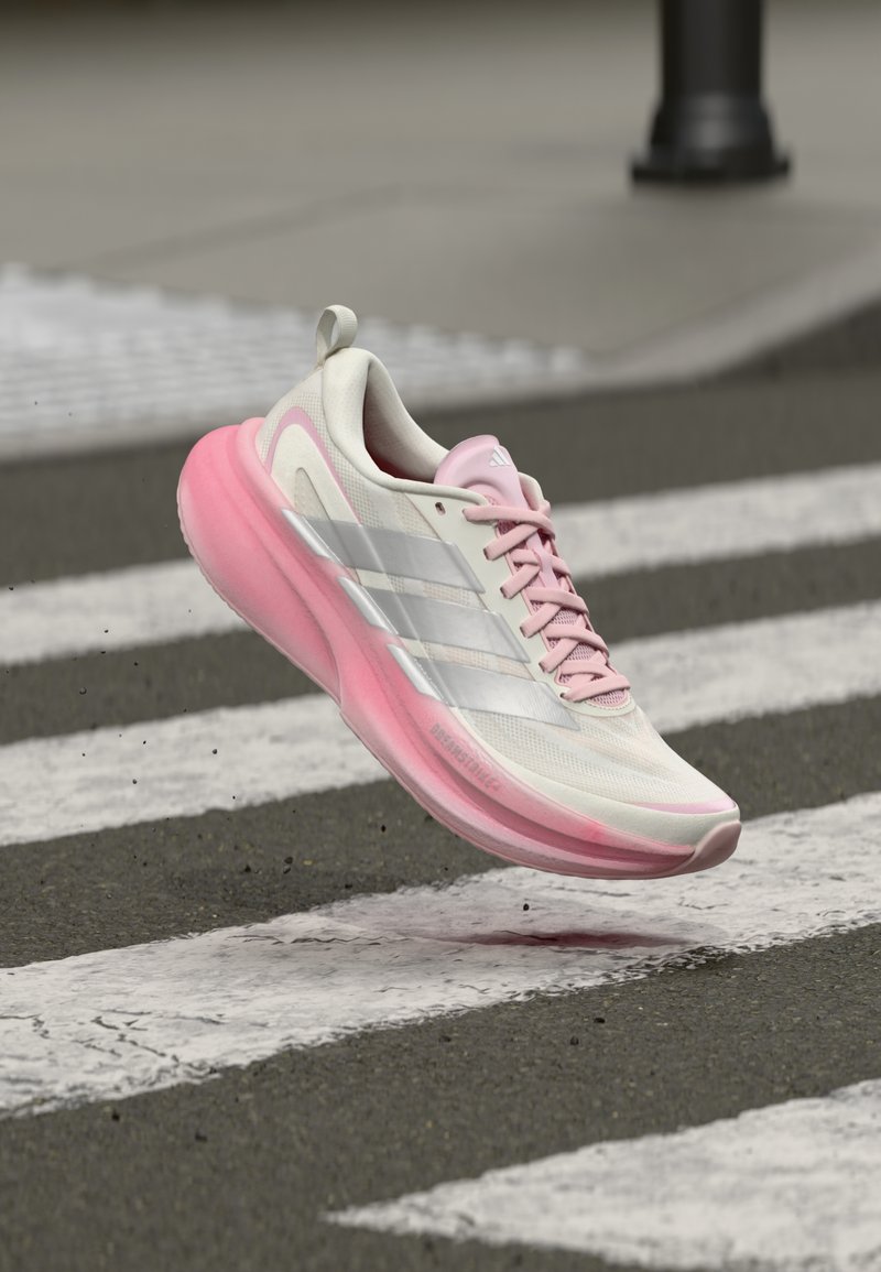 Chaussure de course blanche et rose avec une tige en mesh texturé, trois bandes argentées et une semelle rose rembourrée, capturée en l'air au-dessus d'un passage piéton.