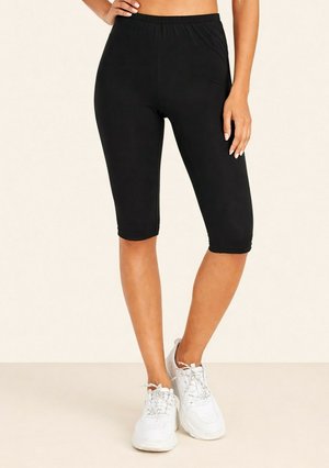 Femme portant des leggings noirs jusqu'aux genoux et des baskets blanches, debout avec une main sur la hanche devant un fond uni.