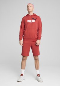 Rød hættetrøje med "PUMA" logo, matchende shorts og hvide sneakers. Glat stof, afslappet pasform og minimalistisk design.