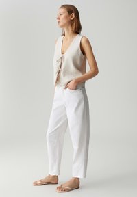 Lichtbeige mouwloze top met een strikfront, gecombineerd met witte taps toelopende broek. Het schoeisel heeft dunne beige bandjes. Minimalistisch ontwerp.
