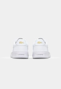 Balti odiniai sportbačiai su plokščiu padu, kuriuose yra auksinė Lacoste logo ir akcentai ant kulno, lygus paviršius ir minimalistinė dizaino forma.