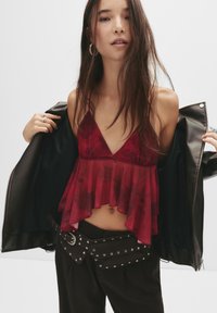 Giovane donna che tiene aperta una giacca di pelle nera, indossando un top rosso trasparente senza maniche, pantaloni neri, una borsa a cintura con borchie e orecchini a cerchio.