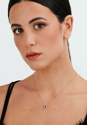 Goldene Kettenhalskette mit einem smaragdgrünen Stein, umgeben von weißen Akzenten, getragen mit einem schwarzen Kleidungsstück, mit glatter Oberfläche.