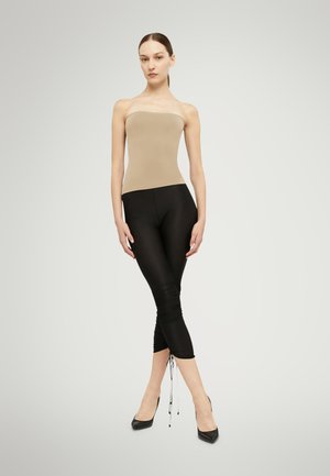 Femme debout devant un fond uni, portant un top beige sans bretelles, des leggings noirs courts avec cordons, et des talons pointus noirs.