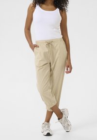 Pantalon de jogging beige rayé avec taille élastique et cordon de serrage, doté de poches latérales et d'ourlets côtelés. Porté avec un débardeur blanc.