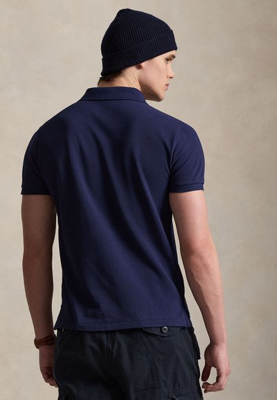 Hombre con camiseta tipo polo azul marino de manga corta, pantalones negros y un gorro de punto oscuro, mirando hacia un lado sobre un fondo liso.