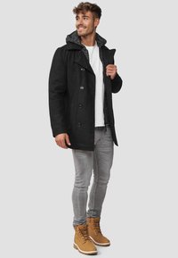 Svart peacoat med knappelukking og grå hette, kombinert med en hvit t-skjorte, grå jeans og brune snøring støvler.