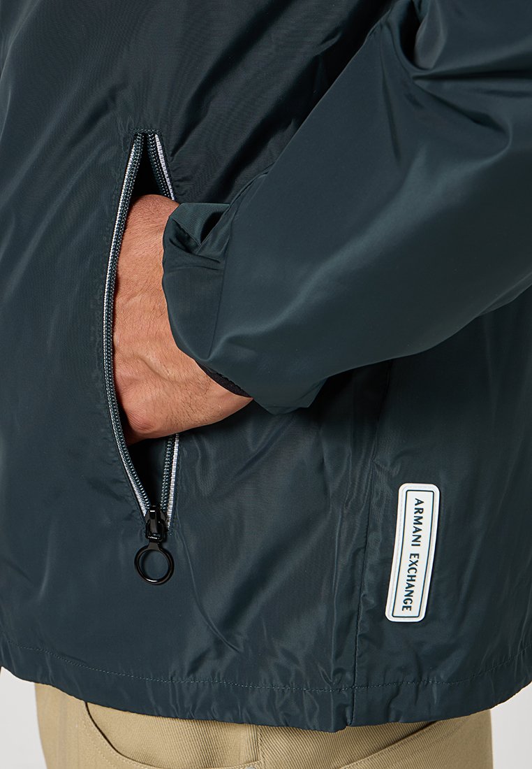 Giacca leggera blu scuro con tasca con zip, polsini elasticizzati e etichetta "Armani Exchange" sul lato. Texture liscia, design casual.