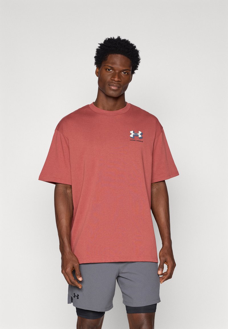 Camiseta de algodón roja con cuello redondo, mangas cortas y un logo azul en el lado izquierdo del pecho, combinada con pantalones cortos deportivos grises.