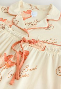 Cream pajama set with orange piping, buttons, drawstring waist, and gingerbread man and dessert illustrations with "Ginger Bread Martini" text.
