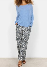 Frau in einem hellblauen Langarmpullover, mit gemusterten, lockeren Hosen in Blau und Beige, und braunen Sandalen, die mit Händen in den Taschen dasteht.