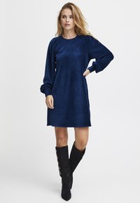 Dunkelblaues Cordkleid mit langen Puffärmeln und einem runden Ausschnitt, kombiniert mit knielangen schwarzen Stiefeln mit dekorativen Akzenten.