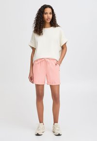Lett stoff kremfarget topp med korte ermer, kombinert med rosa shorts med snøring i livet, rullet kant og sidelommer. Beige joggesko.