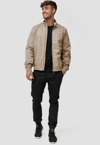 Chaqueta bomber beige con cuello alto, puños acanalados y cierre de cremallera, combinada con pantalones negros ajustados y zapatillas negras.