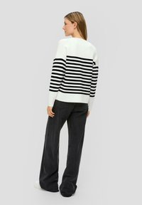Gestreifter Pullover in Schwarz und Weiß mit langen Ärmeln, rundem Halsausschnitt und überschnittenen Schultern, kombiniert mit hochgeschnittenen dunklen Jeans und weißen Turnschuhen.