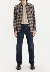 Rutiga flanellskjortor i tan och marinblått, ihop med en beige t-shirt och mörkblå raka jeans. Har två bröstfickor och knapprand.