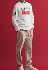Sudadera blanca con el logo "GANT" en navy, combinada con pantalones beige que presentan patrones bordados coloridos, y zapatillas blancas.